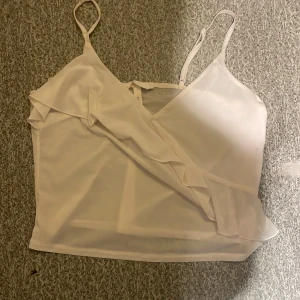 Vit omlottlinne med volang - Säljer ett stilrent vitt omlottlinne från H&M med justerbara axelband och volangdetaljer framtill. Perfekt för varma sommardagar eller som ett lager under en kavaj. Passar till både jeans och kjol. 