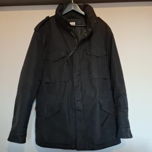 Svart Field jacket från C.P. Company - Säljer en stilren svart jacka från C.P. Company. Jackan har långa ärmar och flera praktiska fickor framtill. Den är perfekt för kyligare dagar och har en klassisk design med dragkedja och knappar.