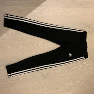 Svarta träningsbyxor från Adidas - Snygga svarta träningsbyxor från Adidas med de klassiska tre vita ränderna längs sidorna. Byxorna är tillverkade i AEROREADY-material som håller dig torr och bekväm under träning. Perfekta för gymmet eller löprundan.
