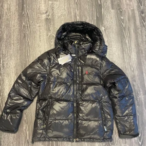 Svart dunjacka från Polo Ralph Lauren - Snygg svart dunjacka från Polo Ralph Lauren med huva och dragkedja. Jackan har en röd logotyp på bröstet och praktiska fickor med dragkedja. Perfekt för kyliga dagar.