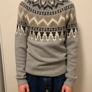 Grå mönstrad stickad tröja - Säljer en grå stickad tröja med ett snyggt geometriskt mönster i svart, beige och vitt. Tröjan har långa ärmar och en bekväm passform, perfekt för kyligare dagar. Den är både stilren och mysig.
