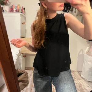 Svart ärmlös topp - Snygg svart ärmlös topp i ett enkelt och lite högre krage. Perfekt för en avslappnad look eller att klä upp med accessoarer. Passar bra till jeans eller kjol.