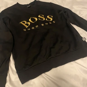 Svart tröja från Hugo Boss - Snygg svart tröja från Hugo Boss med guldigt BOSS-tryck på framsidan. Tröjan har långa ärmar och en klassisk rund halsringning. Perfekt för en stilren look.