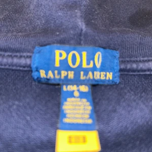 Mörkblå hoodie från Ralph Lauren - Säljer en stilren mörkblå hoodie från Ralph Lauren med dragkedja och klassisk logga på bröstet. Tröjan har en bekväm passform och praktiska fickor framtill. Perfekt för en avslappnad look.