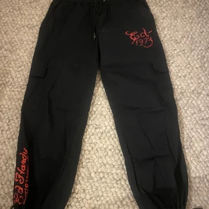 Ed hardy tech byxor - Snygga svarta byxor från Urban Outfitters med ett coolt drakbroderi i flera färger på benet. Byxorna har snörning i midjan och textdetaljer i rött. Perfekta för en avslappnad stil med en unik touch.