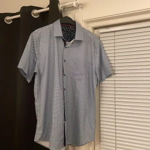Mönstrad kortärmad skjorta - Snygg kortärmad skjorta med ett diskret mönster i blått och vitt. Perfekt för en avslappnad stil. Skjortan har knappar framtill och en klassisk krage. 