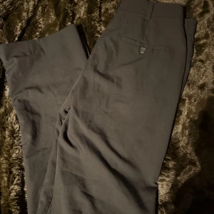 Svarta kostymbyxor från ASOS Design - Snygga svarta kostymbyxor från ASOS Design. De har en klassisk design med raka ben och en knappdetalj vid midjan. Perfekta för en stilren look.