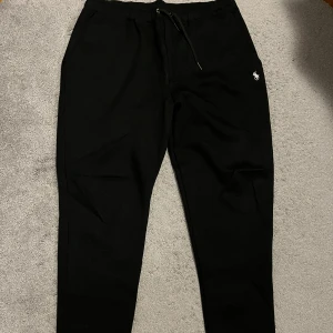 Svarta mjukisbyxor från Ralph Lauren - Snygga svarta mjukisbyxor från Ralph Lauren med en liten broderad logga på sidan. Byxorna har en bekväm passform med snörning i midjan för justerbarhet. Perfekta för en avslappnad stil.