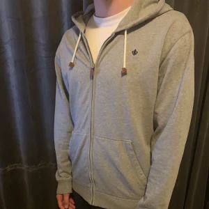 Grå hoodie från Morris - Säljer en stilren grå hoodie från Morris med dragkedja och justerbar huva. Tröjan har en liten broderad logga på bröstet och praktiska fickor framtill. Perfekt för en avslappnad look.