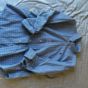 Blå rutig skjorta från Ralph Lauren - Säljer en snygg blå rutig skjorta från Ralph Lauren. Skjortan har långa ärmar och klassiska knappar framtill. Den är tillverkad i bomull och har en broderad logga på bröstet. Perfekt för en stilren look!