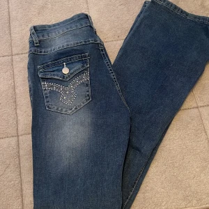 Blå bootcut jeans med broderade fickor - Snygga blå bootcut jeans med broderade detaljer på bakfickorna och knappar. Perfekta för en avslappnad stil med en touch av unik design. Aldrig använt och helt nya de är jätte fina men va lite för stora för mig 