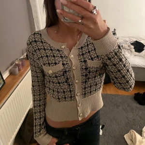 Beige och svart kofta med knappar - Snygg beige och svart kofta med ett rutigt mönster och dekorativa knappar. Koftan har långa ärmar och en croppad passform, perfekt för en trendig look. Storlek S🥰