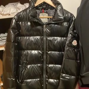 Moncler maya - Säljer en snygg svart pufferjacka från Moncler. Jackan har en svart yta och dragkedja framtill. Den är perfekt för kyligare dagar och har en klassisk Moncler-logga på ärmen. Jackan är dunfylld och certifierad enligt DIST för spårbarhet och integritet.