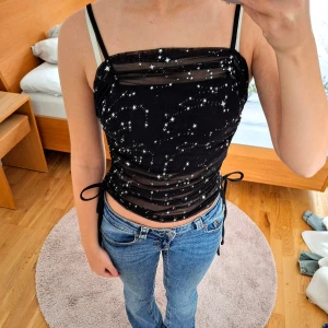 Mesh Star Ruched Top - Svart meshtopp med stjärnmönster och justerbara axelband. Toppen har en åtsittande passform med knytband på sidorna för en extra detalj. Perfekt för en kväll ute eller en speciell tillställning. Använd 1 gång, bortklippt tvättlapp för estetik. Tvättar 30°.🖤