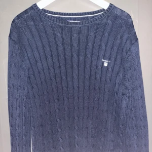 Blå stickad tröja från GANT - Säljer en snygg blå stickad tröja från GANT med kabelstickat mönster. Tröjan har en rund halsringning och långa ärmar. Perfekt för kyligare dagar! Passar även om du har M.