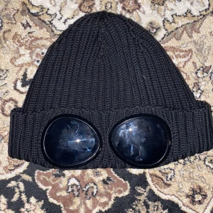 Svart stickad mössa med glasögondetalj från C.P. Company - Snygg svart stickad mössa från C.P. Company med unika glasögondetaljer framtill. Perfekt för att ge din outfit en cool touch. Tillverkad i ett mjukt och bekvämt material.
