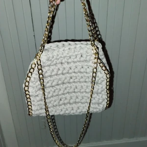 Grå virkad handväska med guldkedja - Säljer en unik grå virkad handväska med en elegant guldkedja. Perfekt för att ge din outfit en personlig touch. Väskan är handgjord och har en mjuk textur.