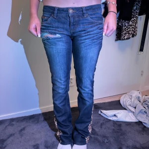 Blå jeans med leoparddetaljer - Snygga blå jeans med unika leoparddetaljer vid bensluten. De har en klassisk femficksdesign och en knappgylf. Perfekta för att ge din outfit en cool twist. Waist:26 längd 32