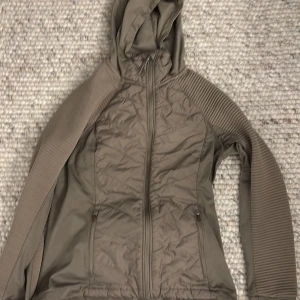 Beige jacka med huva - Säljer en beige jacka med huva och dragkedja. Jackan har långa ärmar med ribbade detaljer och praktiska fickor framtill. Perfekt för kyligare dagar.