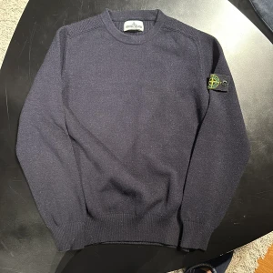 Mörkblå tröja från Stone Island Junior - Säljer en stilren mörkblå tröja från Stone Island Junior. Tröjan har en klassisk design med ribbade muddar och en ikonisk logga på ärmen. Perfekt för en avslappnad och trendig look.