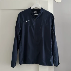 Mörkblå vindjacka från Nike - Säljer en stilren mörkblå vindjacka från Nike med dragkedja framtill. Perfekt för sportiga aktiviteter eller som en lätt ytterjacka. Går även bra till en Casual outfit. Storlek ser ni på bilden men den stämmer inte riktigt in, skulle säga att jackan passar S/M i herr storlek. Hör av er vid intresse