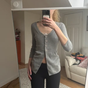 Grå kofta med knappar - Snygg grå kofta med trekvartsärm och stora knappar framtill. Den har en figurnära passform och är perfekt för lager-på-lager-stil. Koftan är i ett mjukt material som ger en bekväm känsla.