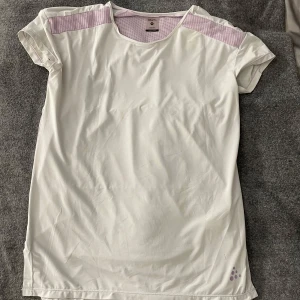  Lila t-shirt från Craft - Säljer en vit t-shirt från Craft med lila meshdetaljer på axlar och rygg. Perfekt för träning med sitt lätta och andningsbara material. Kortärmad design med rund halsringning.