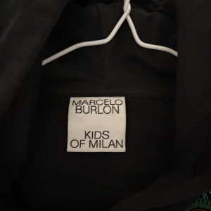 Svart hoodie med luva från Marcelo Burlon Kids of Milan - Väldigt populär Snygg svart hoodie köpt för 3 år sedan och använd 6-7 gånger från Marcelo Burlon Kids of Milan med färgglada broderade detaljer i grönt och gult runt axlarna. Tröjan har en huva och är tillverkad i en blandning av bomull och polyester. 