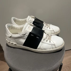 Valentino open - Snygga vita Valentino sneakers med en bred svart rem över snörningen. Skorna har en stilren design med diskreta nitar runt hälen. Perfekta för en trendig look. Box, dustbag, nya snören medföjer. Har order bekräftelse och kvitto. Storlek: 45,5. Har 45 i storlek och passar mig bra.