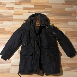 Svart parkajacka från Parajumpers - Säljer en svart parkajacka från Parajumpers med flera praktiska fickor och en varm huva. Jackan har en dragkedja och knappar framtill samt en snygg patch på ärmen. Perfekt för kyliga dagar. Pm för mer bilder. 