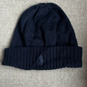 Mörkblå stickad mössa från Ralph Lauren - Snygg svart stickad mössa från Ralph Lauren med ribbad kant och broderad logga framtill. Perfekt för kyliga dagar och en stilren look.