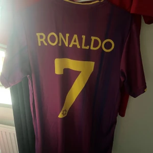 Ronaldo tröja - Säljer en lila fotbollströja från Dune med gult tryck av 'Ronaldo' och nummer 7. Tröjan har korta ärmar och klubbmärke på bröstet. Perfekt för fotbollsfans! I storlek M