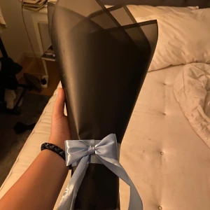 Bukett med 7 blå rosor - En elegant bukett med konstgjorda blå rosor, inslagna i svart papper och dekorerad med ett ljusblått band. Perfekt som en stilren och unik present eller dekoration.