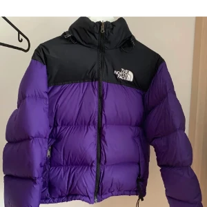 The North Face Jacka - En snygg och varm jacka i lila färg med svart huva. Jackan har en dragkedja framtill och två fickor på sidorna. Den har en snodd i midjan för att justera passformen.  Den är jätteskön och håller dig varm med dess puff. Jackan har två små hål