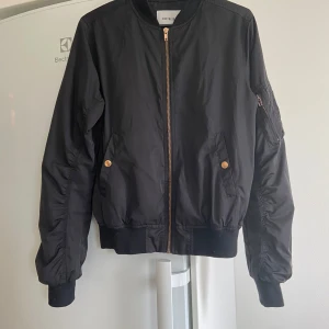 Svart bomberjacka från Size & Needle - Säljer en snygg svart bomberjacka från Size & Needle. Jackan har en dragkedja och praktiska fickor i Gulddetaljer. Den är långärmad och har en ribbad krage samt muddar vid ärmslut och nederkant. Perfekt nu inför hösten. Jackan är bra skick. 