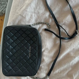 Svart quiltad axelväska från Michael Kors - Säljer en snygg svart axelväska från Michael Kors. Väskan är quiltad och har en stilren design med en silverfärgad dragkedja. Den har en justerbar axelrem och är perfekt för både vardag och fest. Väskan är i bra skick och redo för nya äventyr!