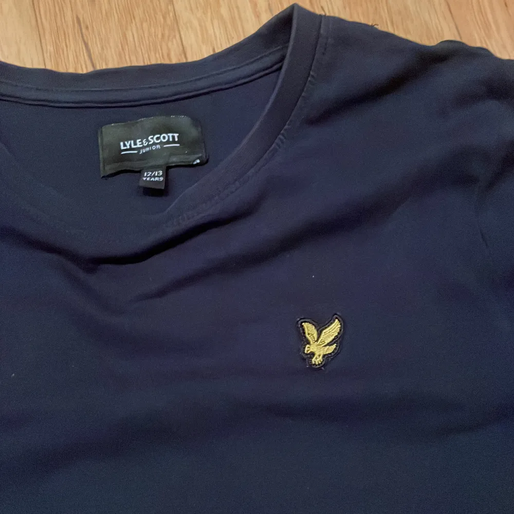 Säljer en snygg mörkblå t-shirt från Lyle & Scott i juniorstorlek 12/13 år. Den har en klassisk design med ett Lyle & Scott märke på bröst. Perfekt för vardagsbruk och passar till det mesta. T-shirten är i bra skick och gjord av mjukt bomullsmaterial. T-shirts.