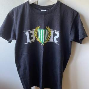 Säljer en svart Bajen t-shirt av siffrorna 1312 med en  grön och vit sköld omgiven av en guldfärgad lagerkrans. T-shirten är kortärmad och perfekt för vardagsbruk. Passar bra till jeans eller shorts!