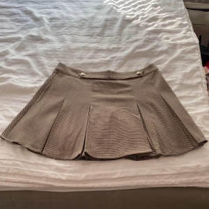 Rutig kjol från H&M - Säljer en superfin rutig kjol från H&M i storlek M. Kjolen har ett klassiskt rutmönster i beige och brunt, med dekorativa knappar framtill. Den är perfekt för både vardag och fest och ger en snygg och stilren look. Kjolen är i bra skick och redo för en ny ägare!