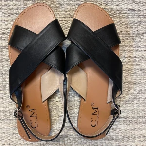 *NY*Svarta Sandaler stl 40 Fr CM Paris  - Snygga svarta sandaler i konstläder från C.M. Paris. Sandalerna har en enkel design med korsade band över foten och en justerbar rem runt hälen.  Stl 40 (innermått 25,4 cm).  Finns ett pytteskav (se bild 5) på ena sidan av en sko, syns knappt. 