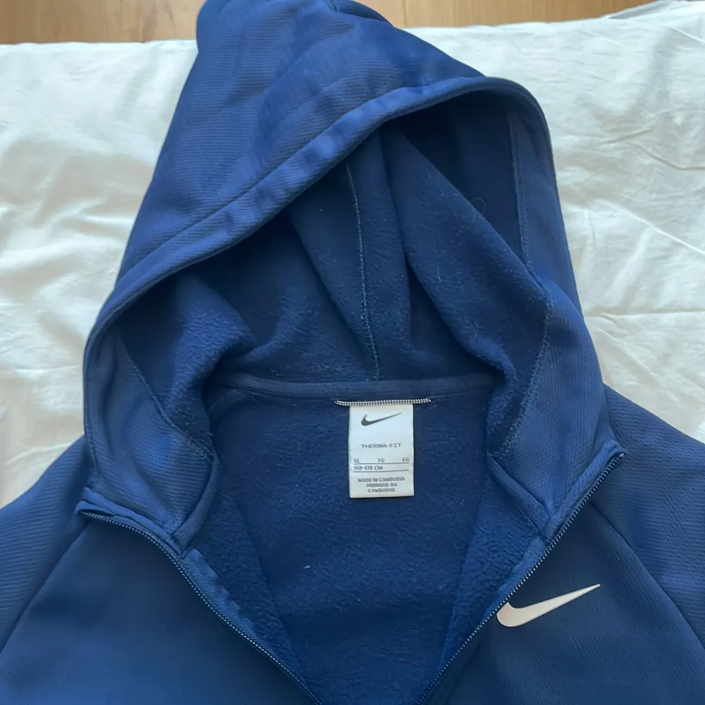 Säljer en mörkblå hoodie från Nike i storlek barn XL. Den är gjord av mjukt och varmt Therma-FIT-material, perfekt för kyliga dagar. Hoodien har en dragkedja framtill och en klassisk Nike-logga på bröstet. Den är i bra skick och redo för nya äventyr!. Tröjor & Koftor.