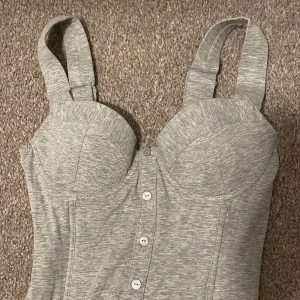 Grå korsett från Lindex - Säljer en snygg grå korsett från Lindex. Den har knappar framtill och breda axelband för extra stöd. Baksidan har en elastisk detalj för bättre passform. Perfekt för en stilren look!
