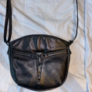 Svart axelväska från Fancy - Säljer en stilren svart axelväska från märket Fancy. Väskan är gjord i syntetmaterial och har en praktisk dragkedja upptill. Perfekt för vardagsbruk eller en kväll ute. Den har en justerbar axelrem för bästa passform.