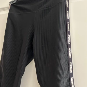 Svarta leggings från Stronger - Säljer ett par svarta leggings från Stronger. De har en ribbad textur och en vit logga längs sidan. Perfekta för träning eller en avslappnad dag. Superbekväma och stilrena!