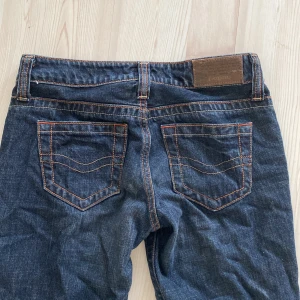 Mörkblå bootcut jeans från C-Seventeen - Säljer ett par snygga mörkblå bootcut jeans från C-Seventeen. De har en klassisk femficksdesign med orange sömmar som ger en cool kontrast. Jeansen är i storlek 27x33 och passar perfekt för en avslappnad men stilren look. 
