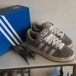 Grå Adidas campus sneakers - Snygga grå sneakers från Adidas med klassiska tre ränder i vitt på sidan. All og medföljer. Skorna har en stilren design med snörning och en bekväm sula. Perfekta för en avslappnad stil. Hör av dig vid funderingar och pris kan diskuteras vid snabb affär mvh Malte 