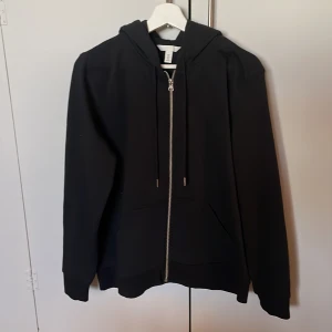 Svart hoodie från H&M - Säljer en stilren svart hoodie från H&M med dragkedja och justerbar huva. Perfekt för en avslappnad look. Den har långa ärmar och praktiska fickor framtill. Perfekt för dig dom är 170 lång ungefär.👍