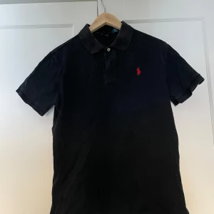 Svart pikétröja från Ralph Lauren - Snygg svart pikétröja från Ralph Lauren med klassisk krage och knappslå. Den har en röd broderad logotyp på bröstet och är i en custom fit passform. Perfekt för en stilren look. Den är lite urtvättad
