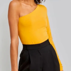 Gul oneshoulder topp från NA-KD - Säljer en snygg gul/orange oneshoulder body från NAKD. Perfekt för en somrig look eller en utekväll. Toppen är i ett mjukt material och har en tight passform. Använd ett par gånger men i bra skick!