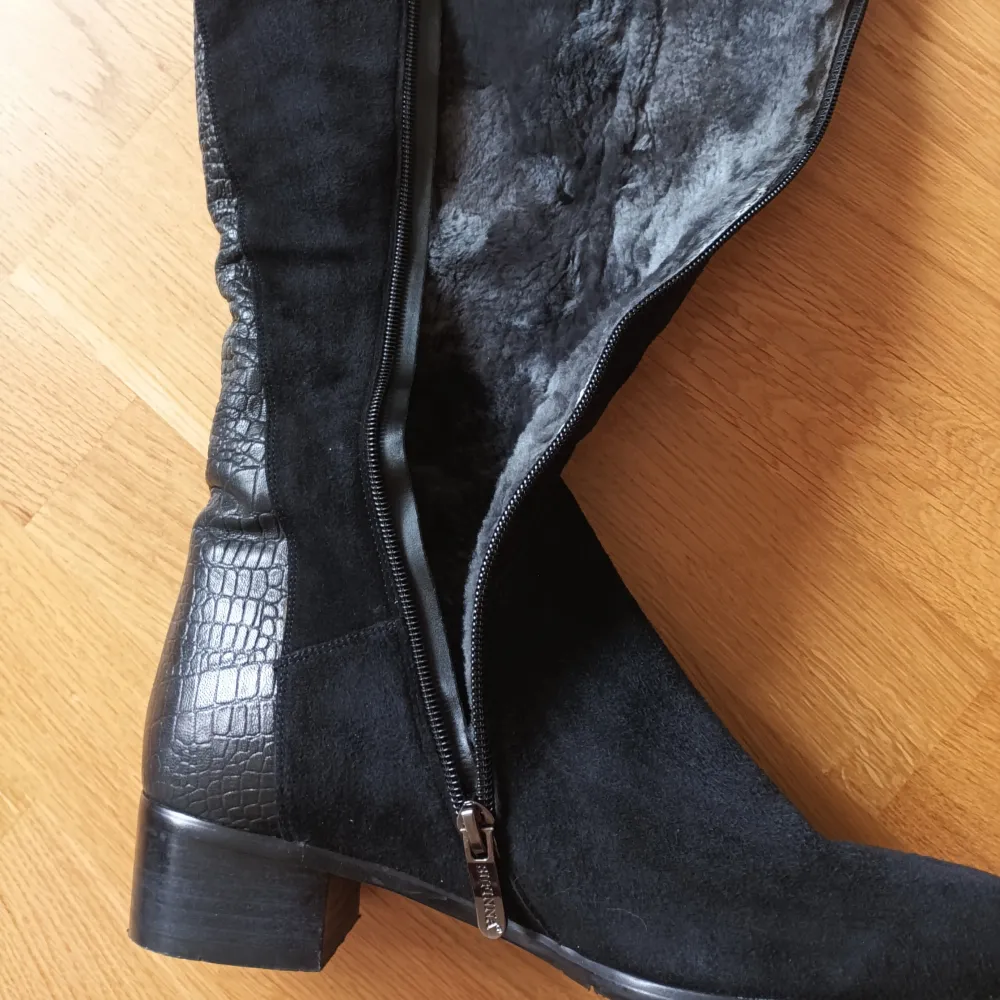 Snygga svarta stövlar i mocka med krokodilmönstrade detaljer och dragkedja på sidan.vinterstövlar i äkta läder med äkta päls inuti. Perfekta för en stilren look. De har en låg klack och är knähöga, vilket ger en elegant touch. . Kengät.